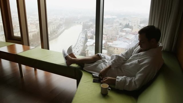 Young man enjoying great cityscape view at luxury apartments and recieving a call on a cellphone sitting on a sofa by window