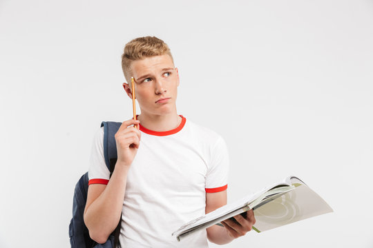 Image of sad smarty guy wearing backpack thinking and touching temple with pen while studying and holding textbooks in hand, isolated over white background