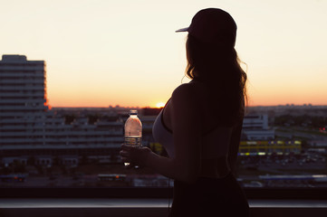 Young woman with bottle water after jogging on the sunset backgrounds. Top view on the city