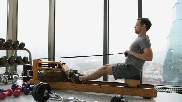 A young fit man trains on a rowing machine in a gym - view from the side