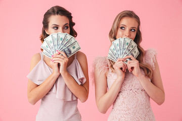 Two successful girls 20s in stylish dresses covering face with fan of money 100 dollar banknotes, isolated over pink background