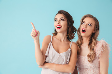 Two beautiful girls 20s wearing party outfit pointing index fingers upward at copyspace with excited faces, isolated over blue background