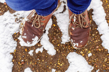 female legs in blue jeans and old brown leather boots stand on a slightly snow-covered stone path