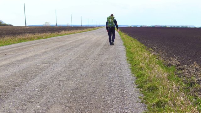 A young tourist walks along a dirt road along the fields with a backpack on his shoulders. Strong wind blows.