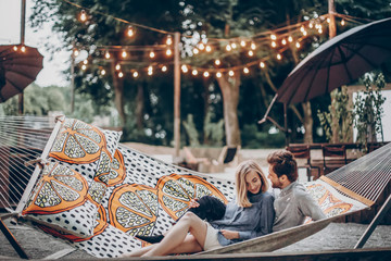 stylish hipster couple cuddling and relaxing in hammock under retro lights in evening summer park....