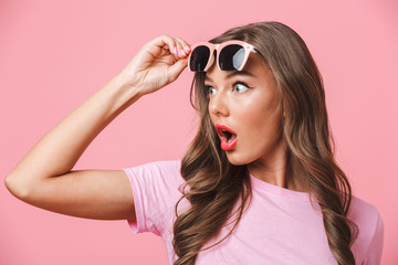Photo closeup of shocked woman 20s looking aside at copyspace with open mouth from under fashionable stylish sunglasses, isolated over pink background