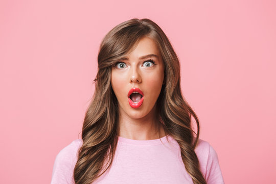 Photo closeup of excited pretty woman with long curly hair in basic t-shirt expressing surprise with open mouth and bulging eyes, isolated over pink background