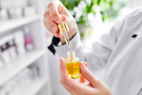 A female doctor-cosmetologist in a white coat holds a bottle of enriched argan oil. Woman with a pipette. Facial care product. Cosmetology room