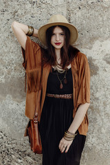 stylish boho woman with jewelry posing at rock wall. beautiful hipster gypsy dressed girl with hat and fringe poncho with sensual look. young girl traveler. fashionable hippie outfit. summer vacation