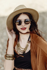 Fototapeta premium beautiful stylish hipster woman portrait in sunglasses holding hat, with windy hair. boho traveler girl in gypsy look, near beach in mountains. atmospheric moment. summer travel.