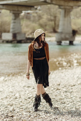 stylish boho gypsy woman in hat, fringe poncho and boots walking on river beach. hipster traveler girl look, summer travel. atmospheric moment. space for text.