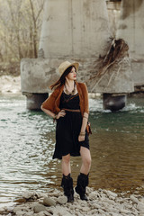 stylish boho woman with jewelry posing at river. beautiful gypsy dressed girl with hat and fringe poncho with sensual look. young girl traveler. fashionable hippie outfit. summer vacation
