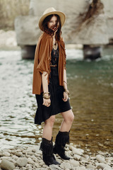 beautiful stylish boho woman smiling, with hat, fringe poncho and boots. girl in gypsy hippie look young traveler posing near river rocks in mountains. sensual look. joyful moment. summer travel