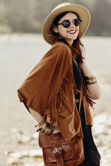 stylish hipster boho woman smiling in sunglasses with hat, leather bag, fringe poncho and...