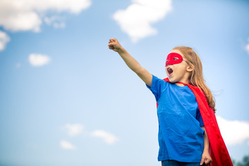 Funny little girl playing power super hero over blue sky background. Superhero concept.