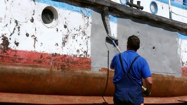 Worker paints metal of old rusty ship propeller at shipyard in port. Process of repair and reconstruction of sea vessel. Technology of manual painting boats. Industry of water transport.