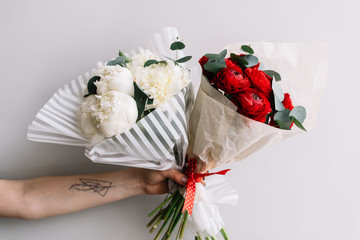 VORONEZH/RUSSIA-02.11.2018: Woman with a tattooed hand holding two colourful fresh blossoming flower bouquets of red ranunculus and eucalyptus and of white  peonies on the grey wall background