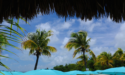 Beach umbrella chairs with blue sky and clouds framed by thatched roof