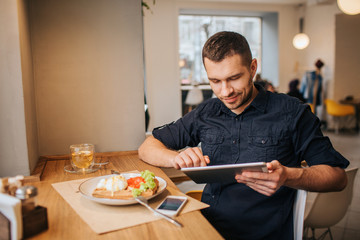 A picture of businessman sitting at table and working. He is using tablet. There is a plate with tasty food and drink on the table. Also there is a phone lying near plate.