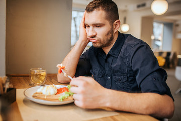 Bored and sad man is sitting at table and cafe. He is holding a piece of vegetable on fork. Man is looking at it and breathing out.
