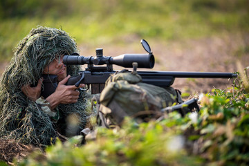 Fototapeta premium Sniper laying on the grass looking through scope at the target in deep forest.