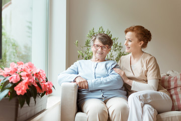 A senior woman with a medical support professional sitting on a sofa while waiting for a geriatric specialist doctor appointment in a private medical clinic. Flowers on a windowsill.