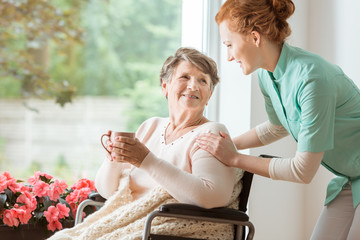 A professional caretaker in uniform helping a geriatric female patient on a wheelchair. Senior...