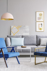 Blue armchair near white table and grey sofa in modern living room interior with poster. Real photo