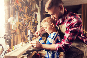 family, carpentry, woodwork and people concept - father and little son with hammer hammering nail...