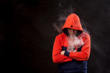 A young man in a orange hood standing in closed posture with lowered head smokes a vape and exhales...