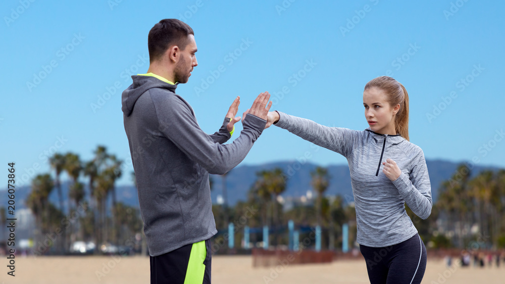 Wall mural fitness, sport, martial arts and people concept - happy woman with personal trainer working on strike over venice beach background in california