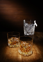 A spotlight on a two crystal glass of scotch whisky with a ice bucket in the backghround, shot on a wooden table top.