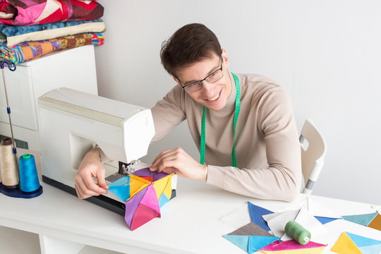 quilting, hobby, passion concept. there is a professional tailor, a young man with adorable smile, who is sitting at his table and stitching patches for feature blanket