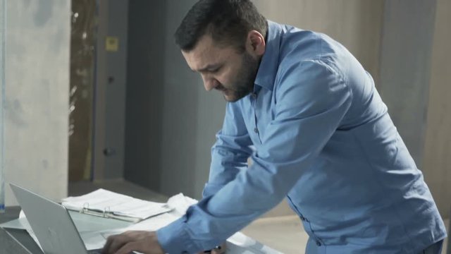 Upset, unhappy man working with laptop and blueprints at his new home
