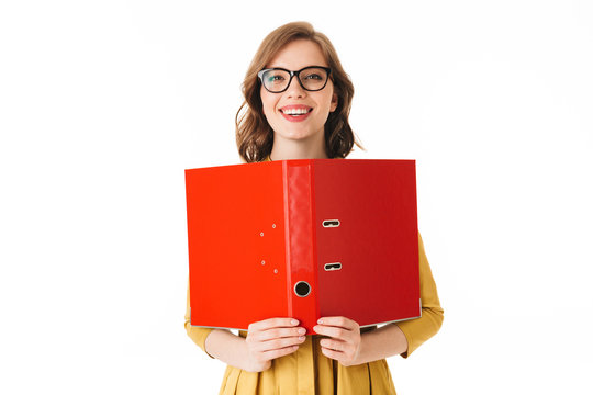 Portrait of young joyful lady in eyeglasses standing with open red folder in hand and happily looking in camera on white background isolated