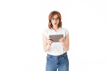 Portrait of young astonished lady in eyeglasses and headphones standing with the tablet in hands on white background isolated