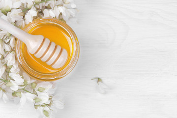 A jar of honey with a wooden spoon for honey and an acacia branch on a white wooden background. top view