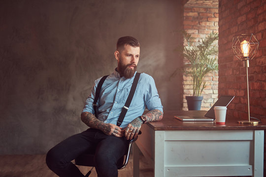 Handsome tattooed hipster in a shirt and suspenders sitting at the desk with a computer, looking out the window in an office with loft interior.