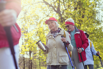 Sunny day. Delighted aged woman holding a thermos bottle while hiking with her friends