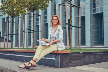 Charming blonde female in modern clothes, studying with a book, sitting on a bench in the park against a skyscraper.