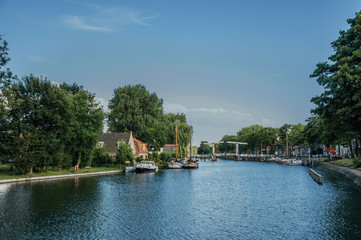 Calm river with grove and bridge, boats moored in the pier of brick houses and sunny blue sky in Weesp. Quiet and pleasant village full of canals and green near Amsterdam. Northern Netherlands.