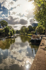 Fototapeta premium Wide canal with cloudy sky, houses and boat moored reflected in water on sunset at Weesp. Quiet and pleasant village full of canals and green near Amsterdam. Northern Netherlands. Retouched photo.