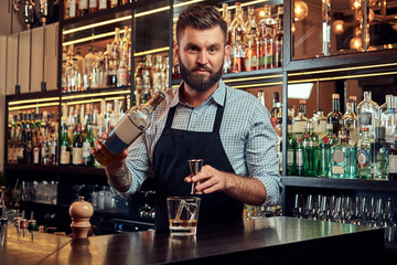 Stylish brutal barman in a shirt and apron makes a cocktail at bar counter background.