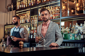 Stylish brutal barman in a shirt and apron makes a cocktail at bar counter background.