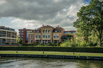 Fancy brick building next to wide tree-lined canal under cloudy sky at sunset in Weesp. Quiet and pleasant village full of canals and green near Amsterdam. Northern Netherlands.