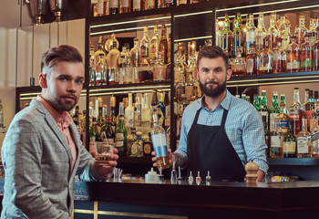 Stylish brutal barman in a shirt and apron makes a cocktail at bar counter background.