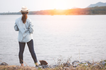 Young traveler woman wear denim shirt standing and looking at lake with chihuahua dog in sunset.