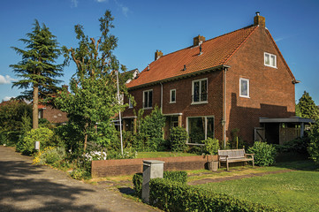 Orange brick house with beautiful and verdant garden in front of alley under blue sky at Weesp. Quiet and pleasant village full of canals and green near Amsterdam. Northern Netherlands.