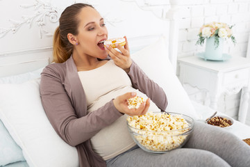 More food. Pleasant pregnant woman resting on bed while chewing junk food