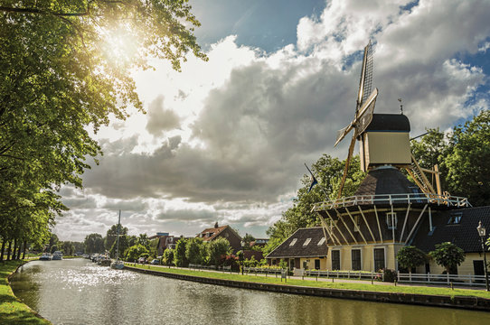 Wide tree-lined canal with windmill and boats and shine of sunset reflected in water at Weesp. Quiet and pleasant village full of canals and green near Amsterdam. Northern Netherlands. Retouched photo